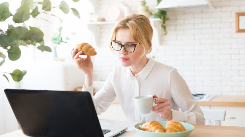Thoughtful business woman using laptop while eating croissant pietu pertrauka darbe