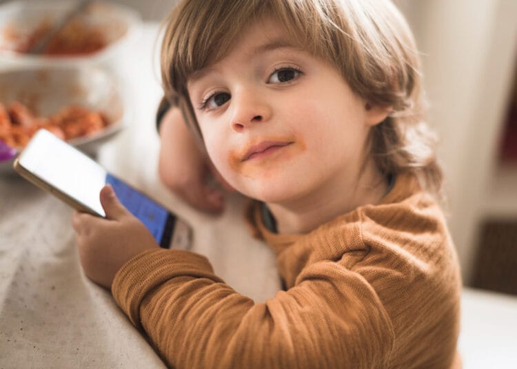 Mielas berniukas prie stalo laikantis telefoną