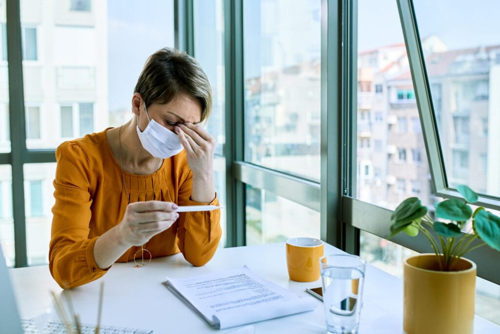 Verslininkė su veido kauke jaučiasi blogai ir matuojasi temperatūrą termometru biure kada mokamas biuletenis