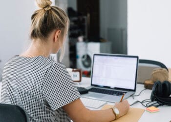 Verslininkės rašančios užrašus ant rudo popieriaus ir nešiojamojo kompiuterio ant stalo vaizdas iš galo