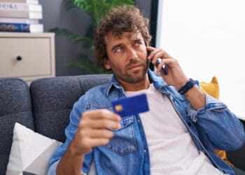 Jaunas ispanakalbis vyras kalba išmaniuoju telefonu ir laiko kredito kortelę sėdėdamas ant sofos namuose kada nereikia moketi vsd