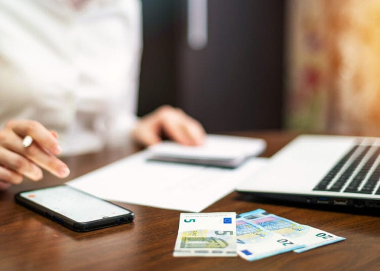 Moteris dirba su finansais ant stalo Nešiojamasis kompiuteris išmanusis telefonas pinigai užrašų knygutė kada taikomas npd