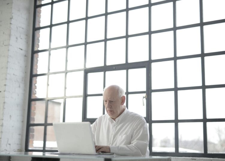 Pensininkas vyras baltais marškiniais sėdintis prie„Macbook kompiuterio dirba